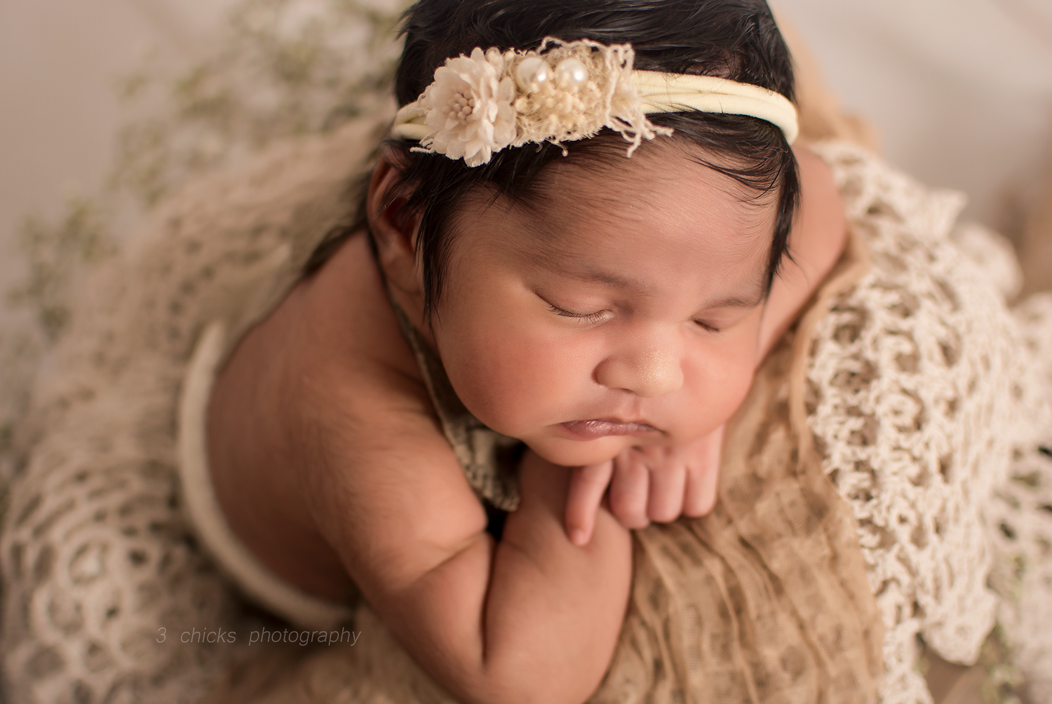 Sleeping newborn baby girl wearing a floral headband, wrapped in cream lace, posed in a cozy basket.