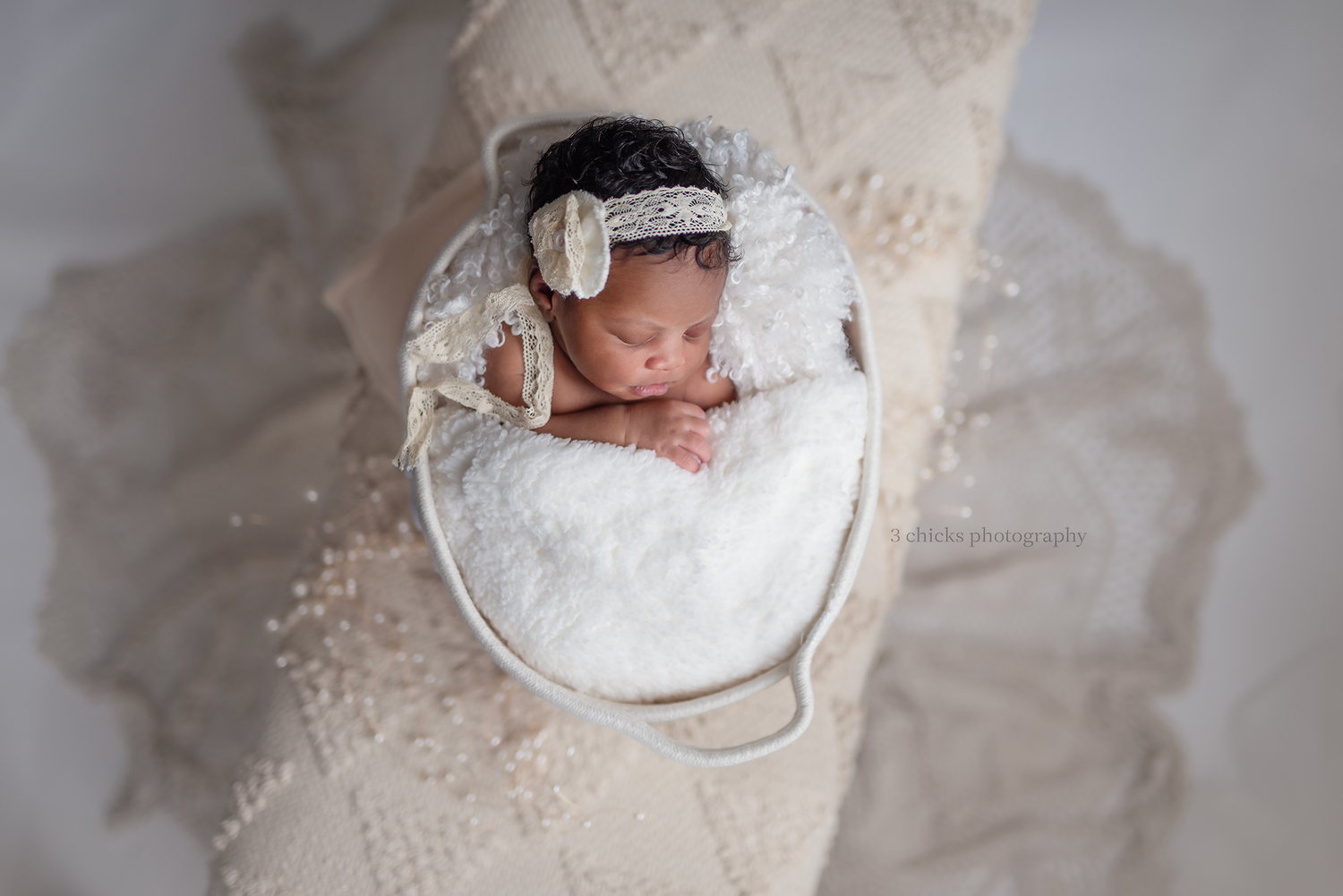 Newborn baby sleeping peacefully in a white bucket with lace headband and white floral outfit on cream backdrop.
