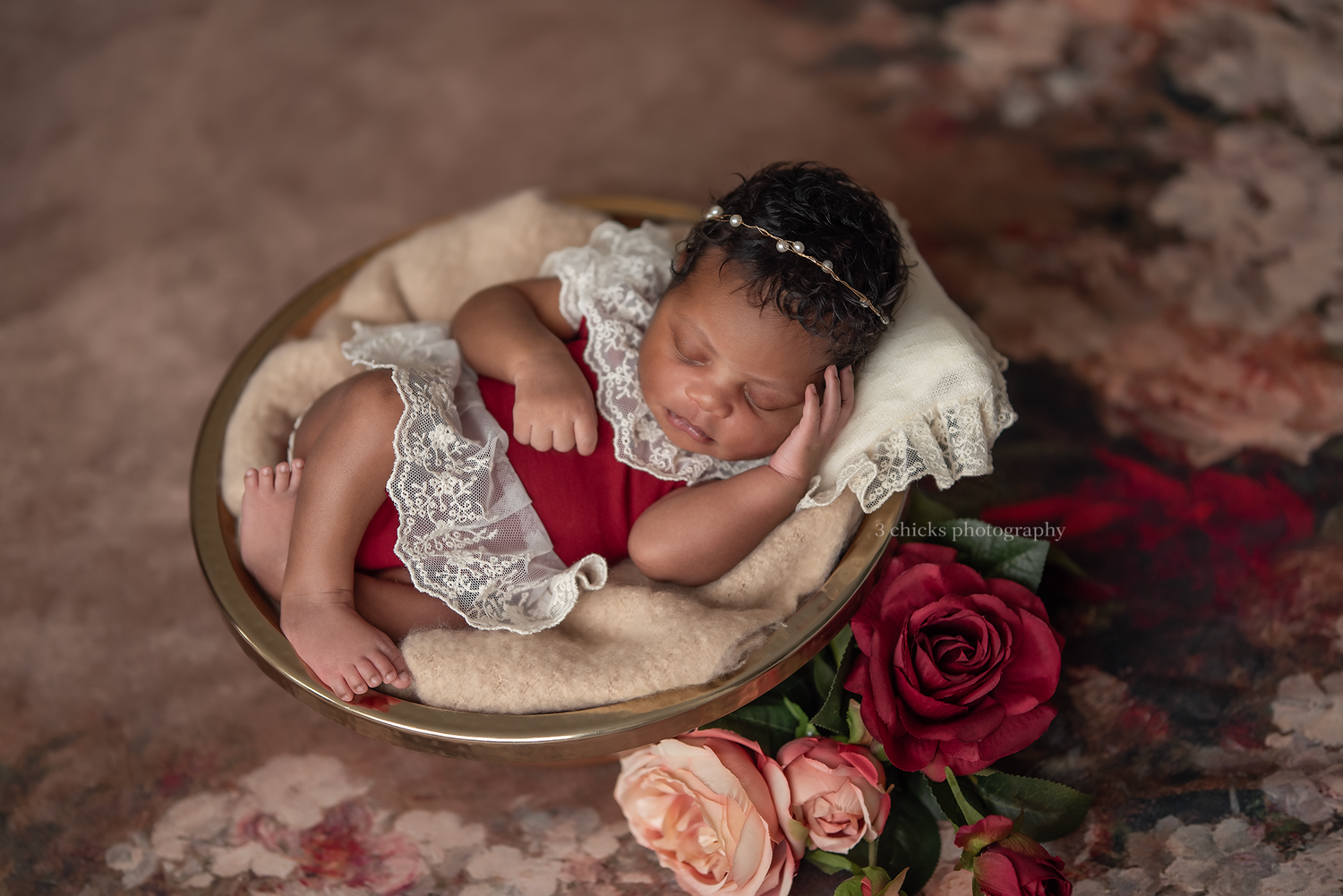 Newborn baby in red lace romper sleeping in gold bowl with roses, wearing pearl headband, on floral backdrop.