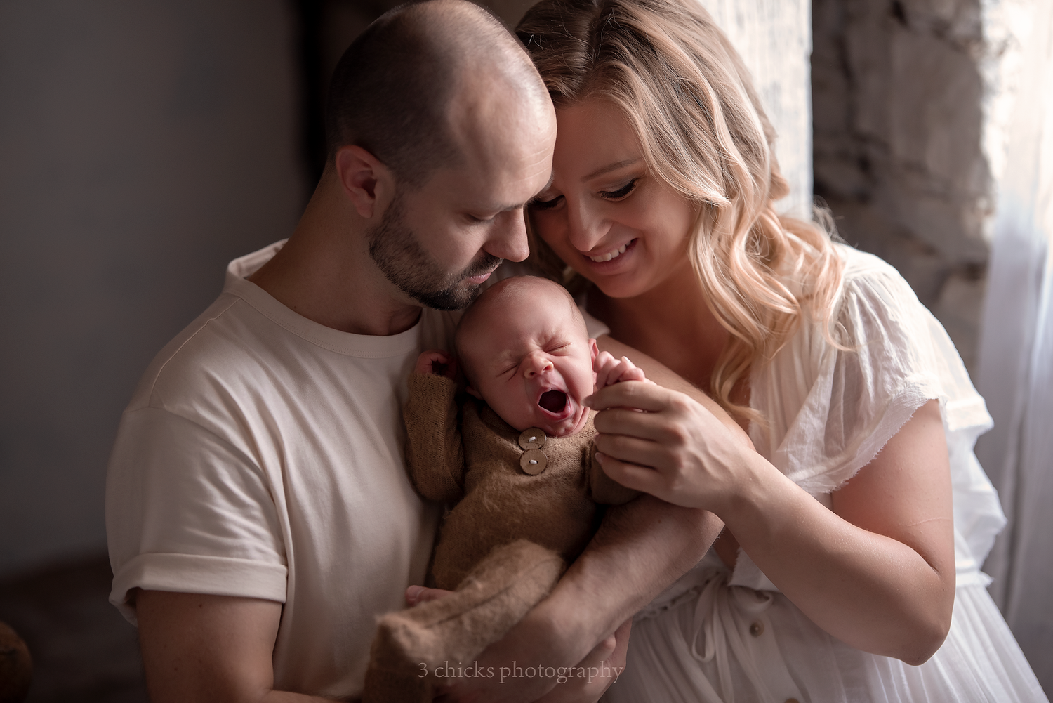 Happy parents smiling and holding their newborn baby wrapped in brown blanket.