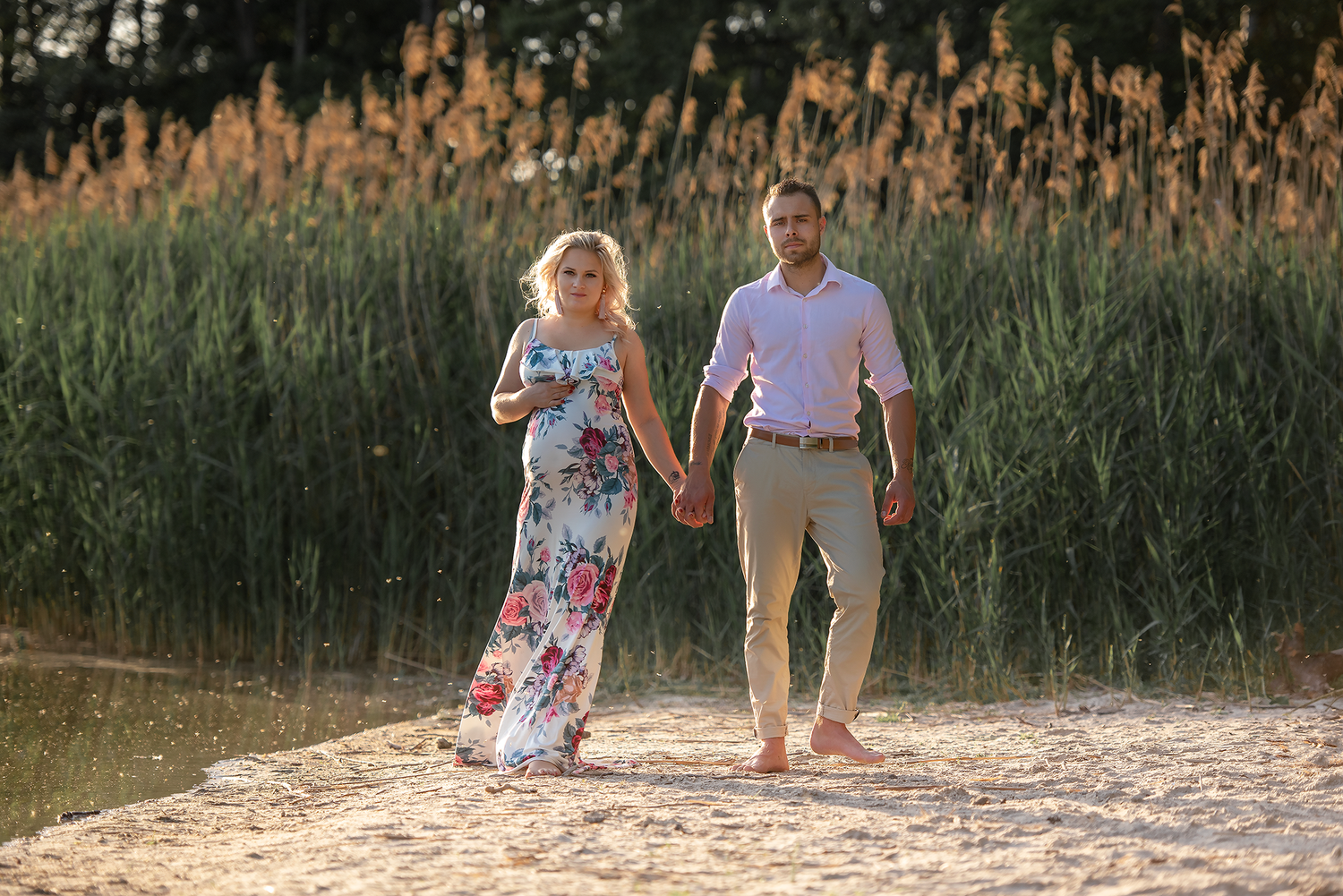 Pregnant woman in floral dress walks hand-in-hand with man on sandy beach near tall green reeds at sunset.