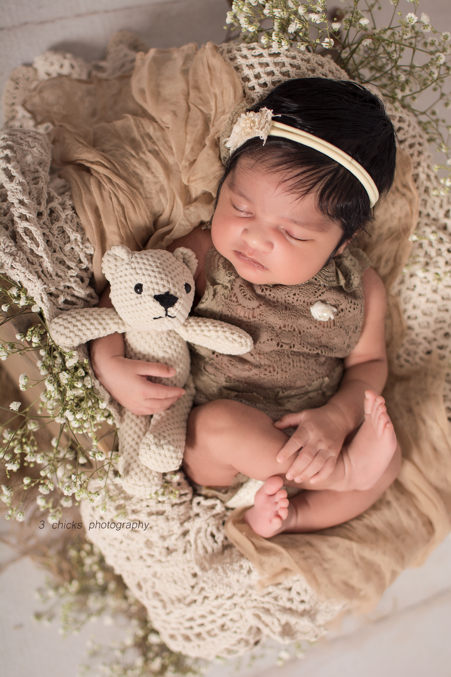 Sleeping newborn baby in brown knit romper holding crochet teddy bear, nestled in basket with lace and baby's breath flowers.