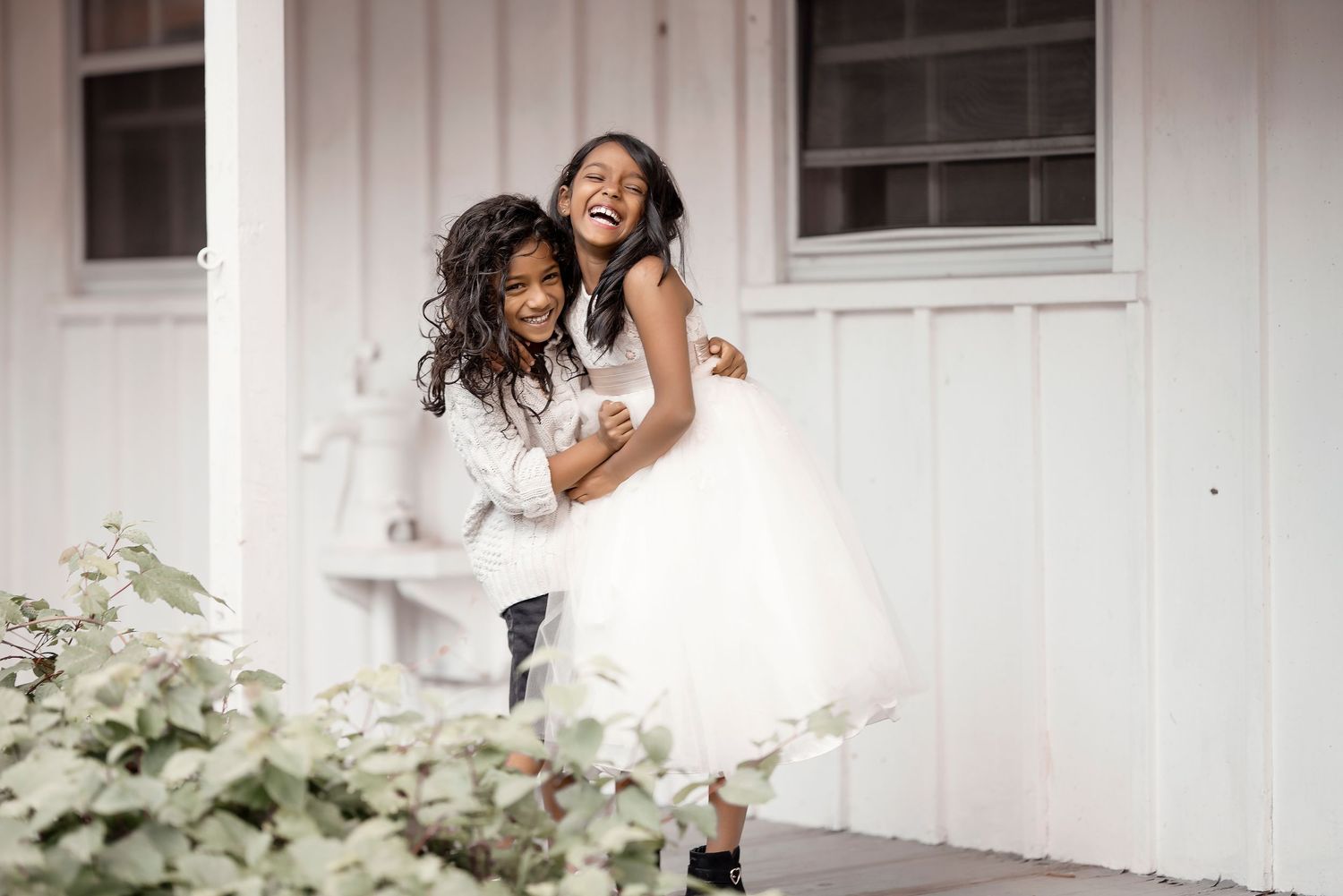 Two young girls in white dresses laugh and embrace joyfully on a porch, capturing a sweet candid moment.