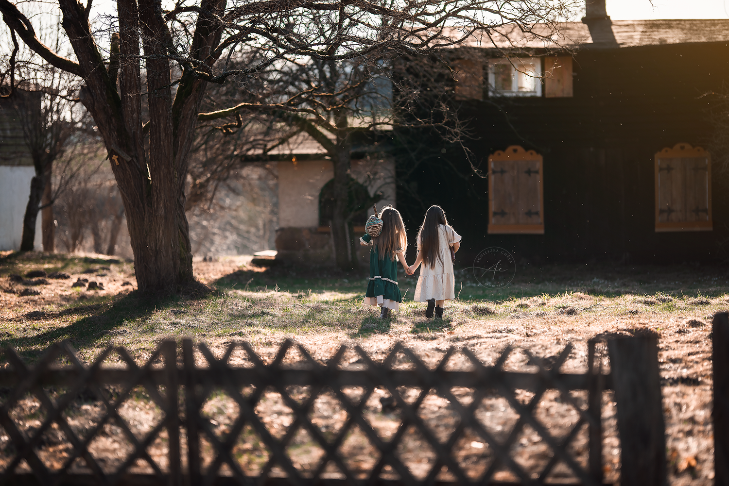 Two young girls in dresses walk toward a rustic house surrounded by trees in warm sunlight.