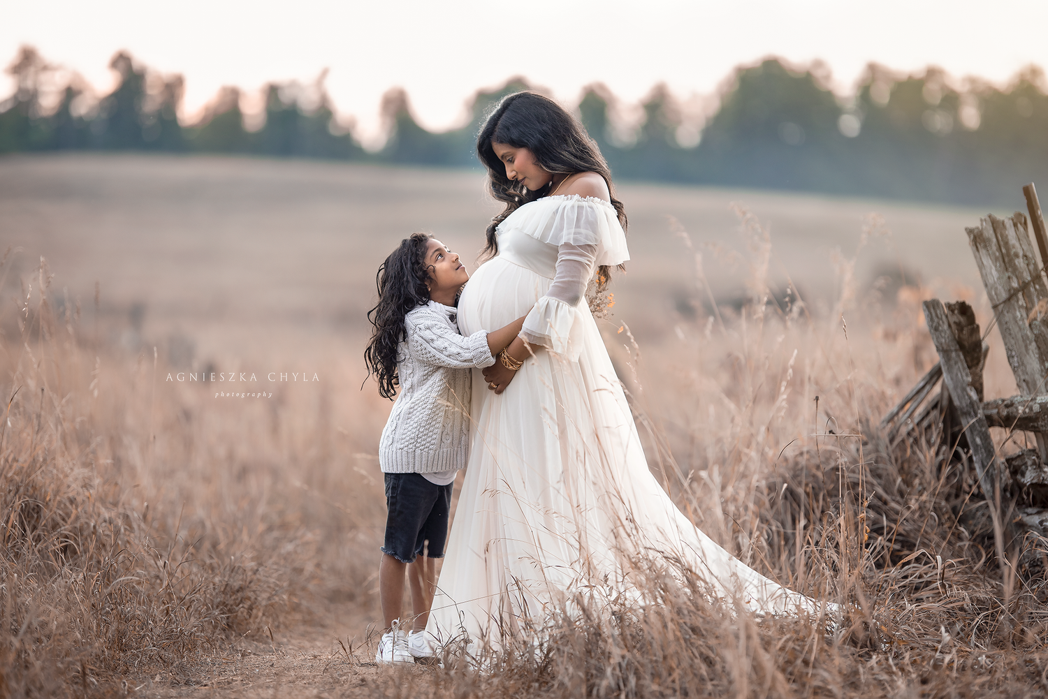 Pregnant woman in white gown embraced by child in sunny field near wooden fence.
