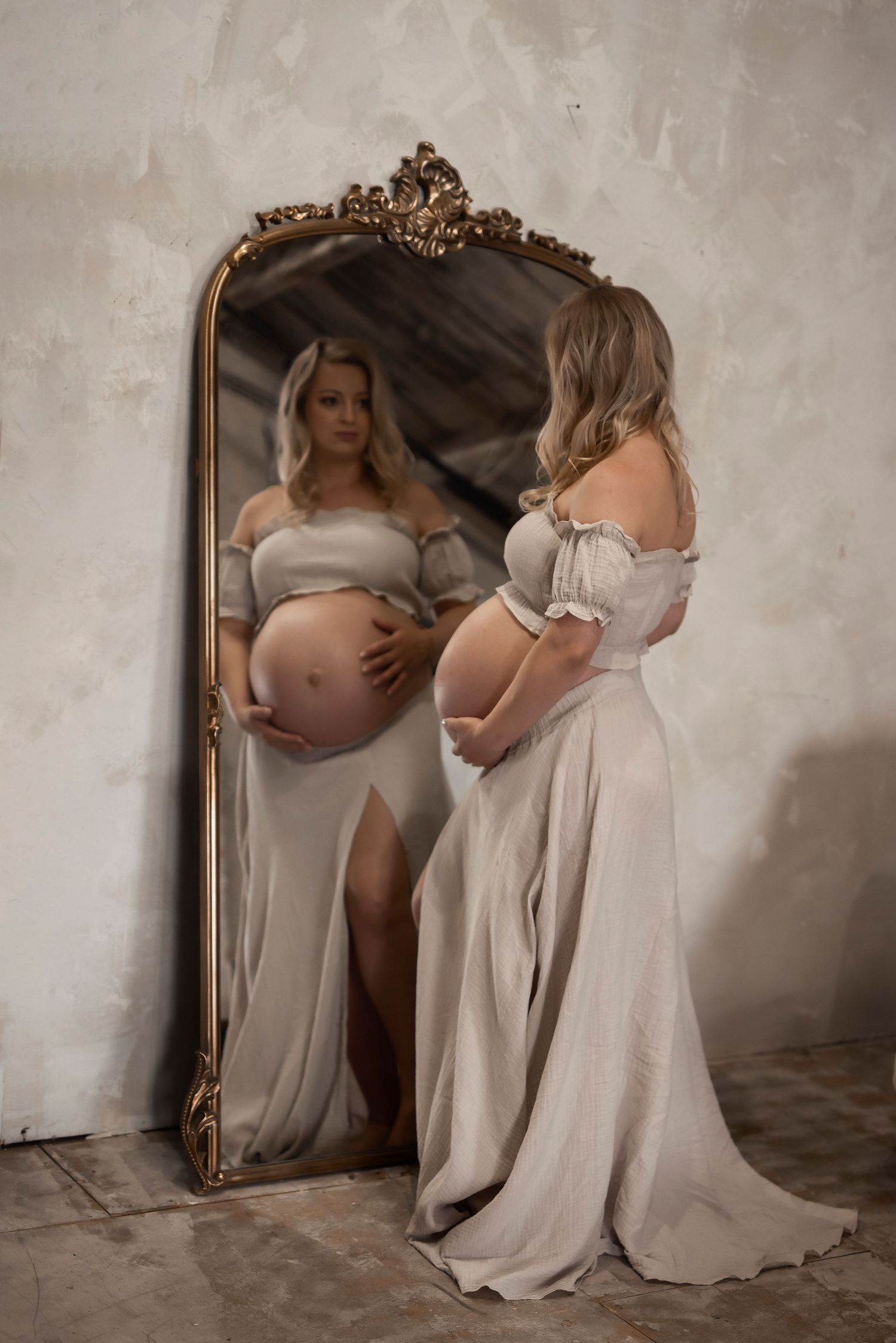 Pregnant woman in elegant gray gown cradling baby bump, reflected in ornate gold mirror against rustic wall.