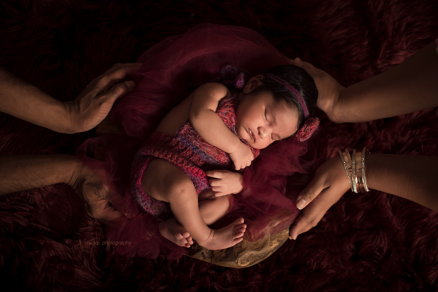 Newborn baby sleeping peacefully in parents hands on red fabric in a dramatic dark photography portrait.