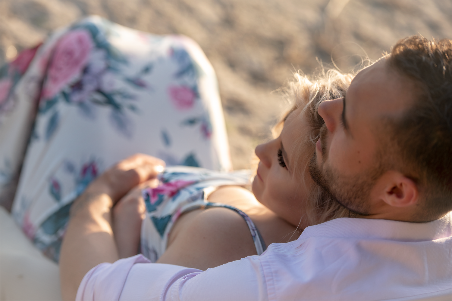 Couple relaxing together on sandy beach, woman in floral dress lying on man's chest in romantic outdoor setting.