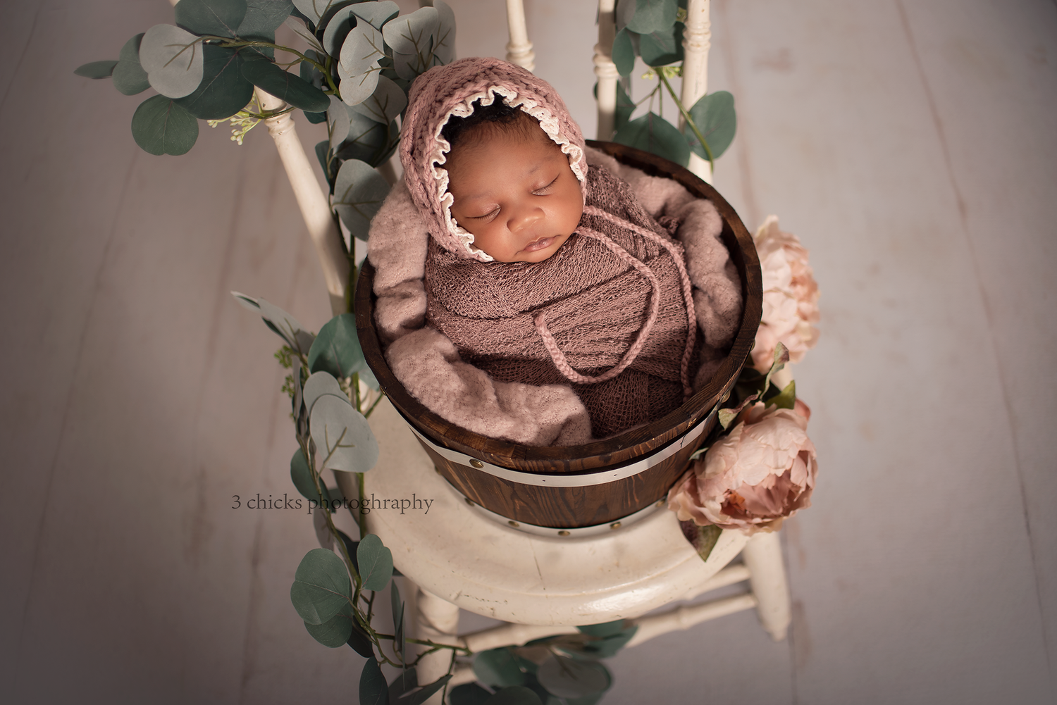 Newborn baby sleeping in brown knit outfit and bonnet inside a wooden bucket on a white chair with greenery.