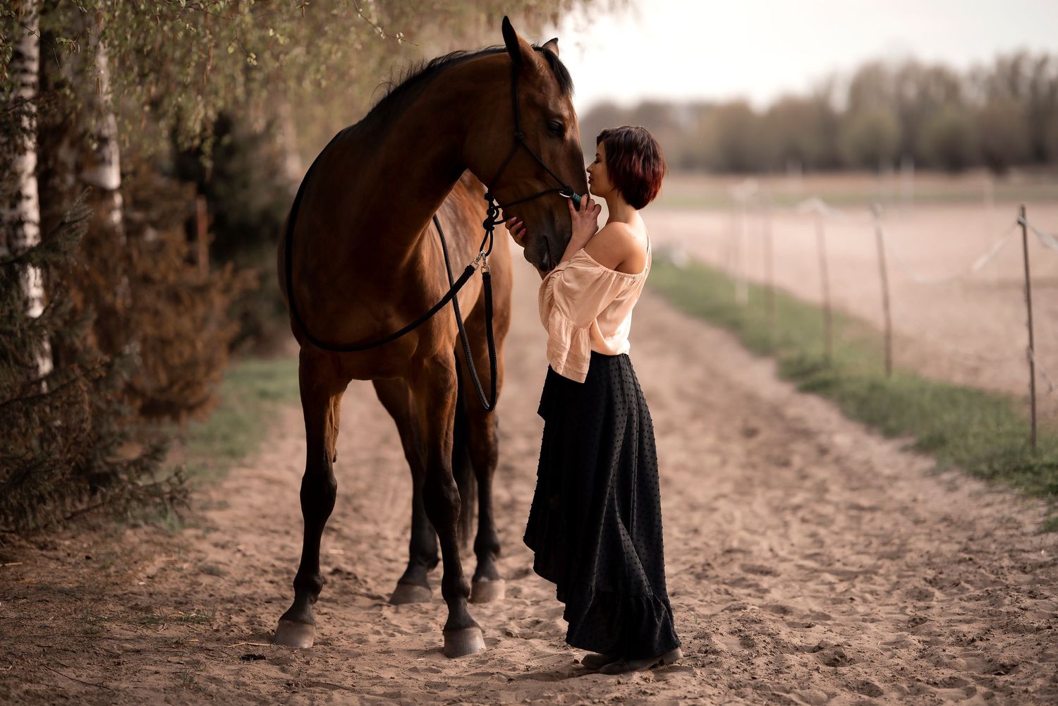 Woman in elegant black skirt and white blouse gently touches a brown horse on a rural path.