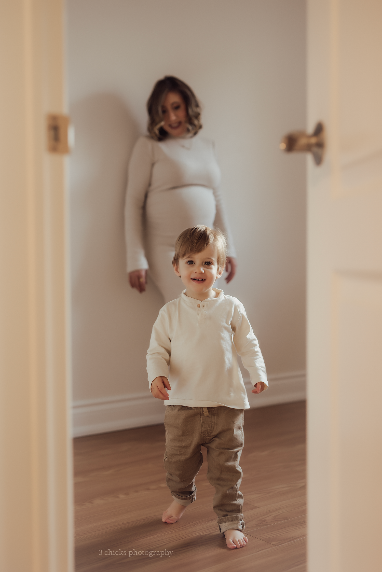 Smiling toddler boy in cream shirt walks toward camera with pregnant mother standing in doorway.