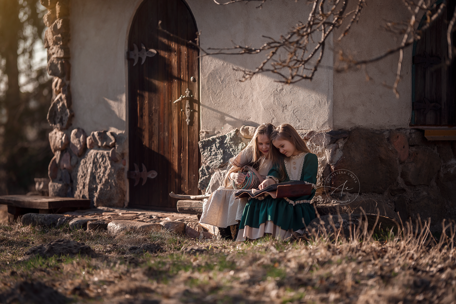Two girls in vintage dresses read a book together on steps of a rustic stone cottage outdoors.
