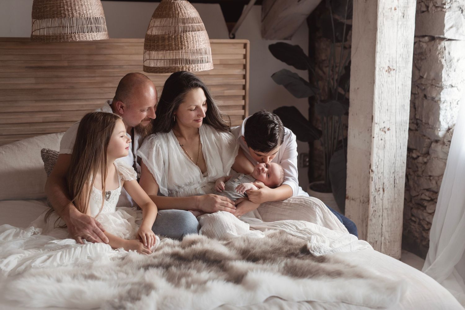Family of five in white outfits sitting on bed together admiring their newborn baby.