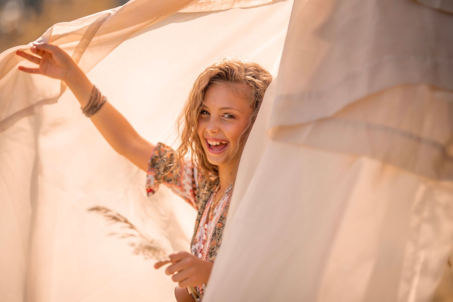 A joyful blonde woman peeks through flowing white fabric in warm golden sunlight, wearing a floral dress.