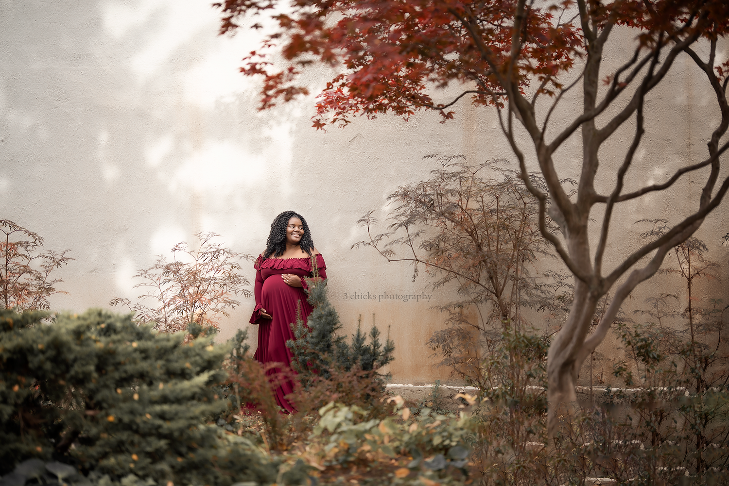 Pregnant woman in red dress standing among autumn trees and shrubs in a misty garden setting.