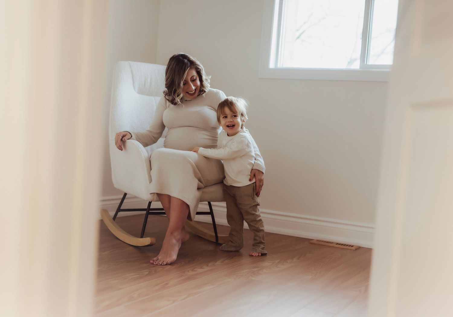 Pregnant woman in white dress sitting in rocking chair with toddler leaning against her in a bright nursery room.