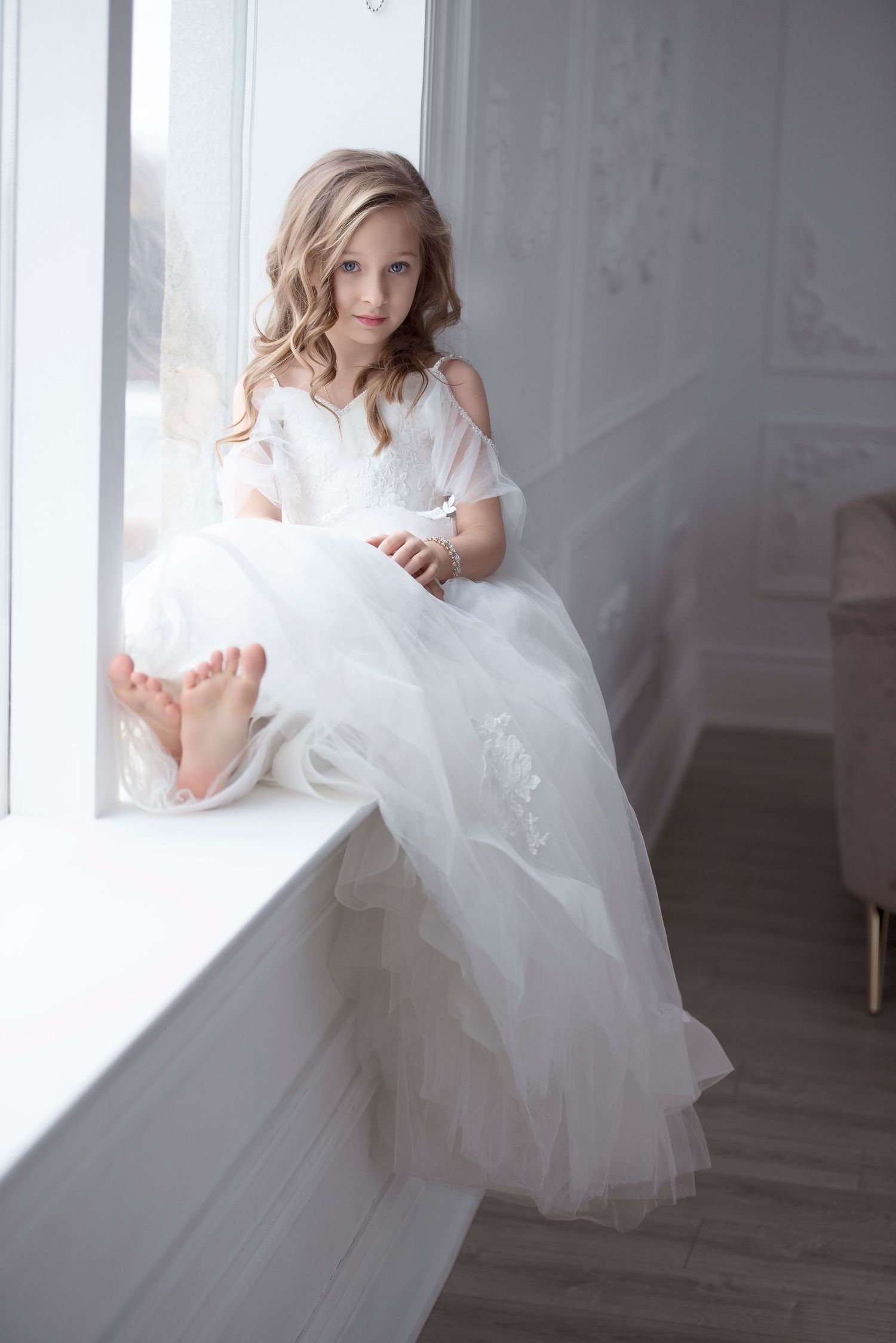 A young girl in a white tulle gown sits barefoot on a bright window ledge.