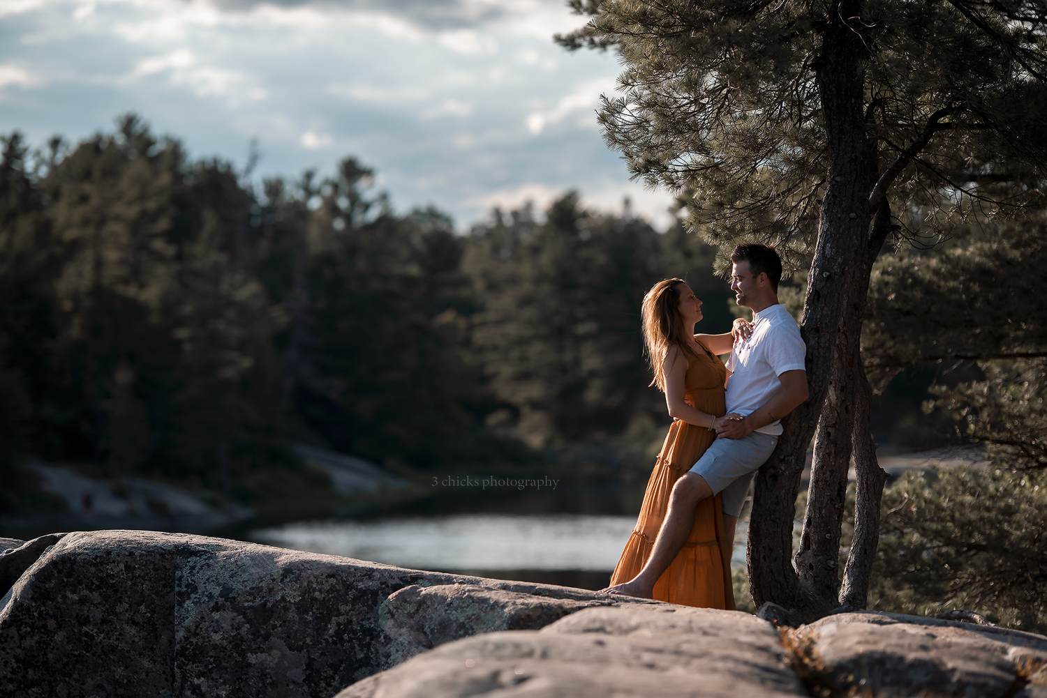 Romantic couple embracing on rocky cliff overlooking a serene forest lake at dusk.