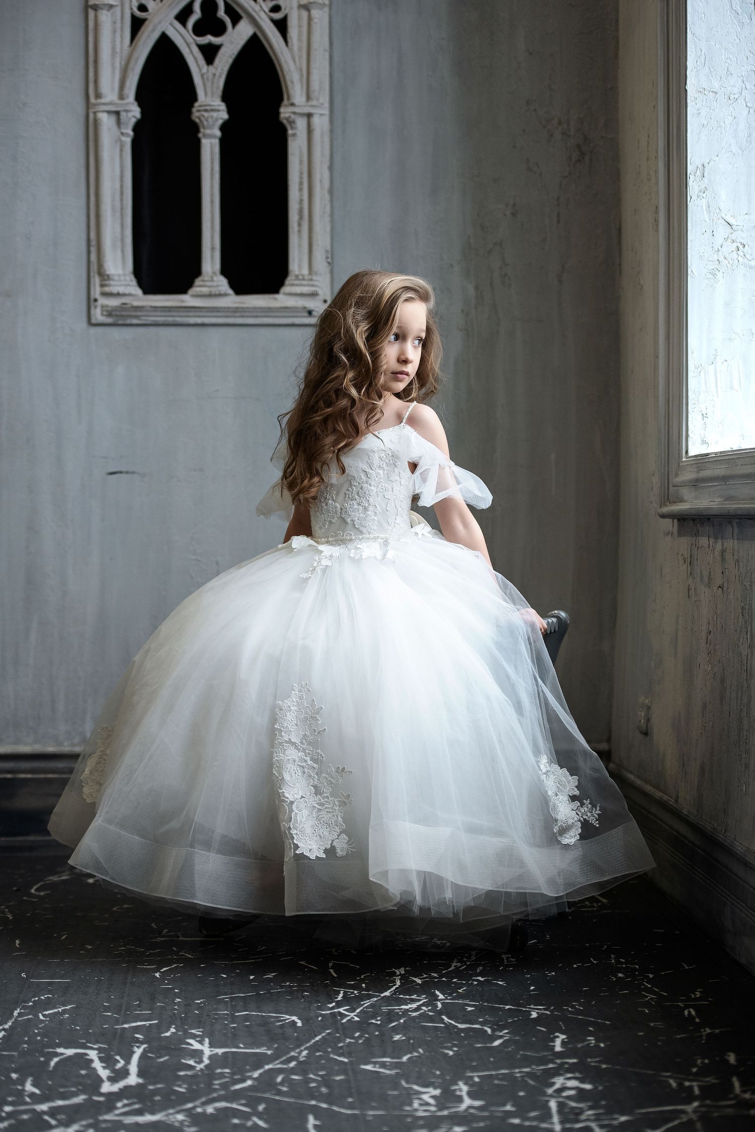 A young girl in a white lace ball gown poses elegantly in a vintage-style room.