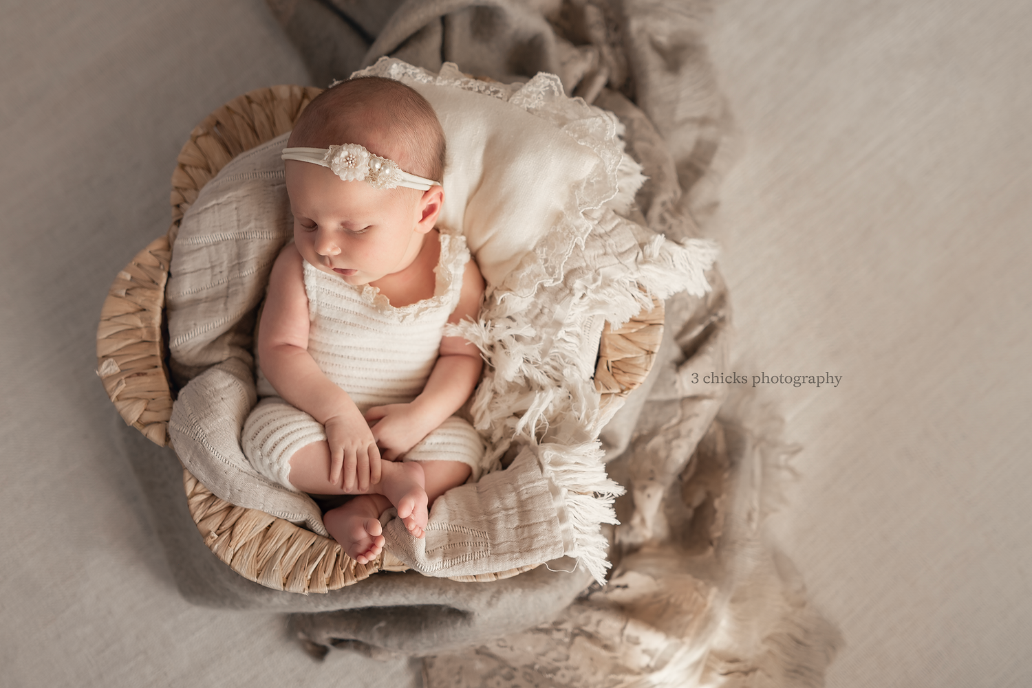 Newborn baby sleeping peacefully in a wicker bowl with cream textiles and a floral headband.