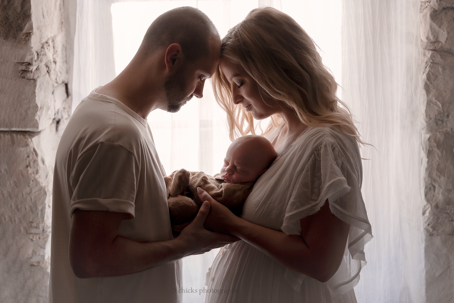 A couple tenderly holds their newborn baby together near a soft-lit window in an intimate family portrait.