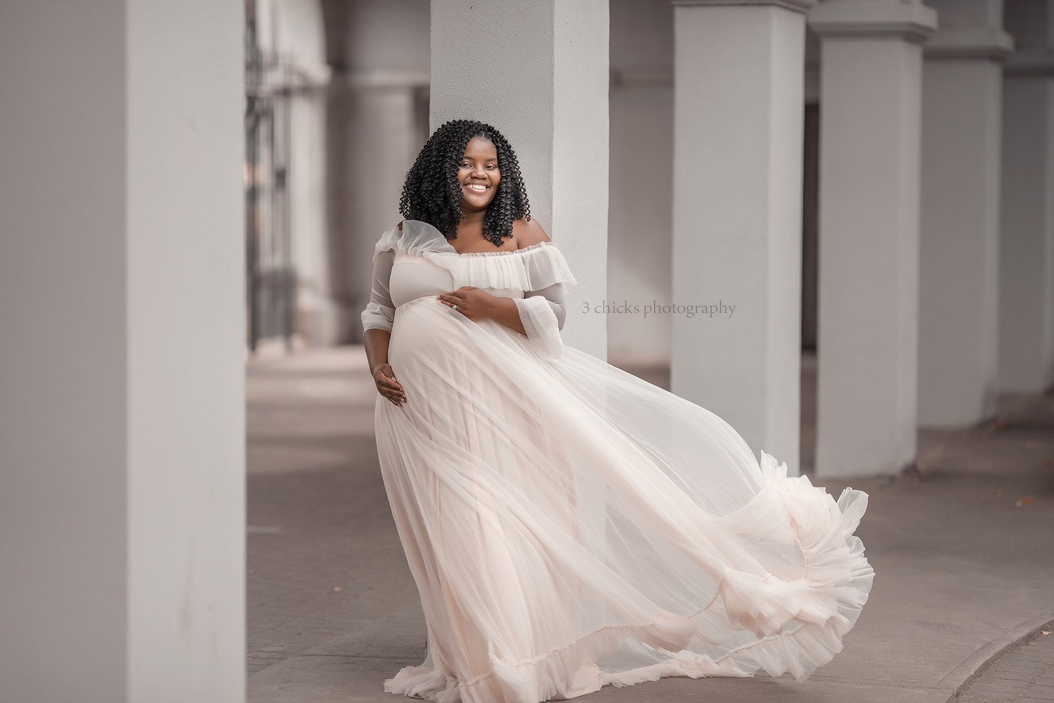 Smiling pregnant woman in flowing white off-shoulder gown poses gracefully between columns in an elegant maternity photo.