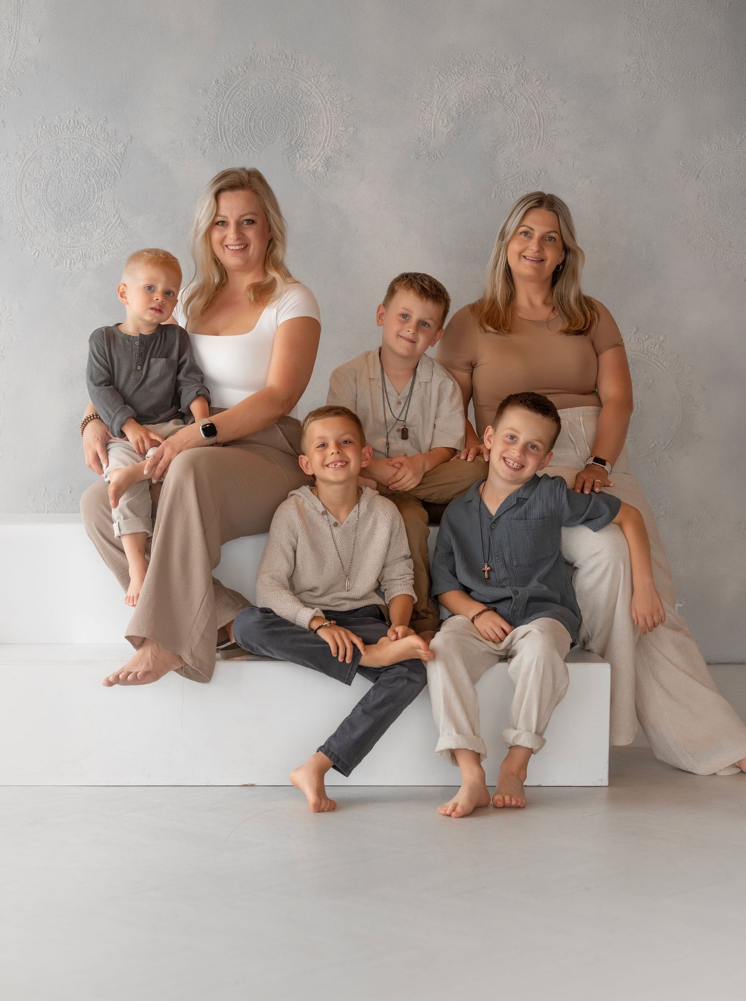 Family portrait of two women and four young boys posing together on a white bench against a gray backdrop.