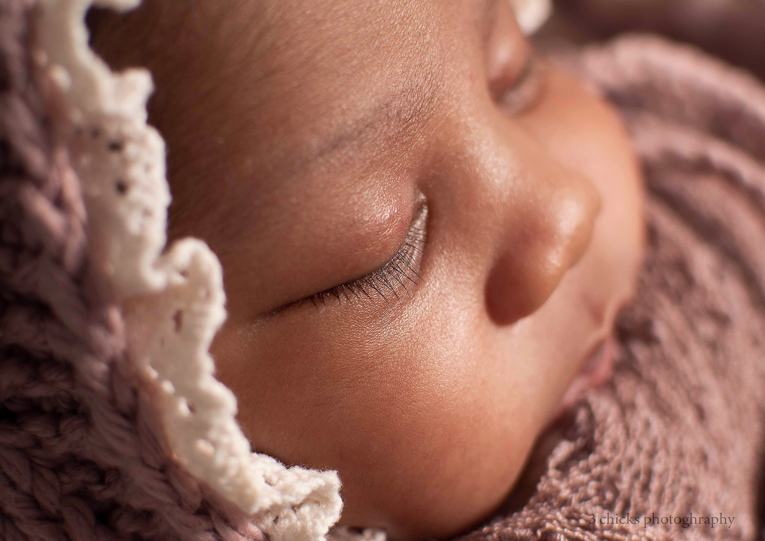 Peaceful sleeping newborn baby wearing a delicate cream crochet bonnet, wrapped in soft mauve knit fabric.