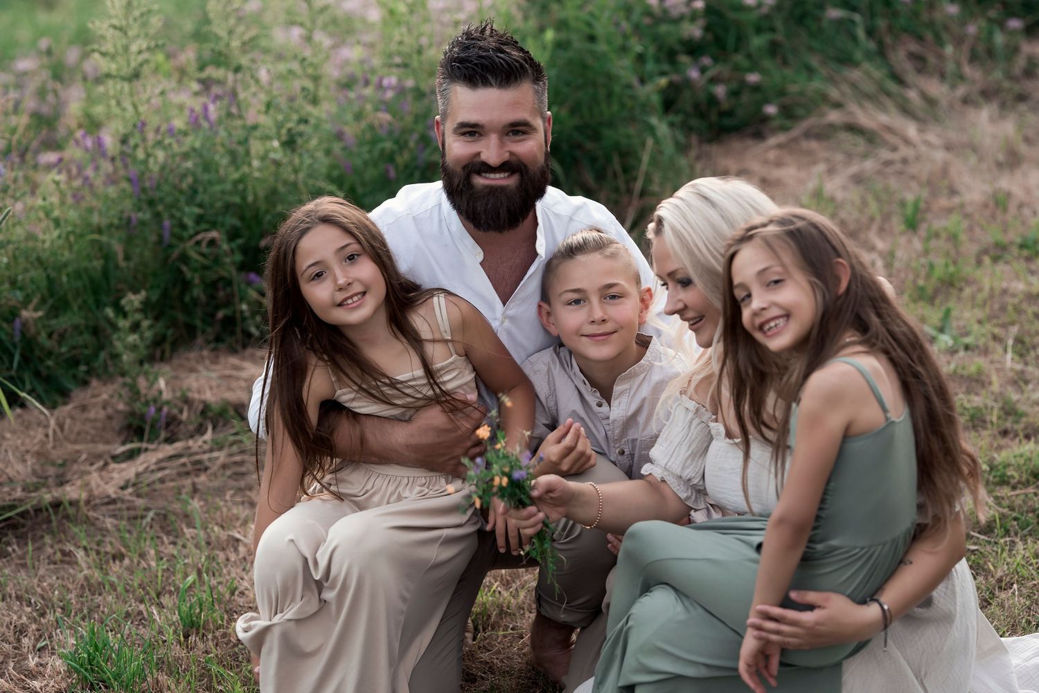 A bearded father poses outdoors with four children in a lush green field during a family portrait session.