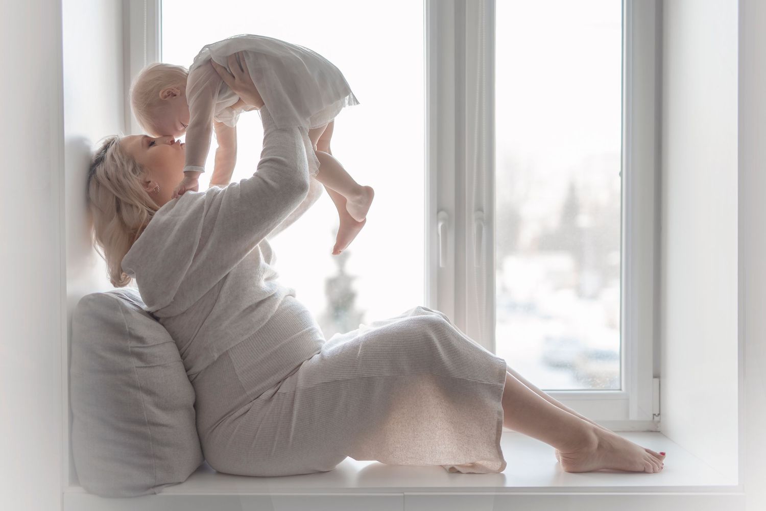 A mother sits on a windowsill lifting her baby in the air, both dressed in white, in a bright, serene room.