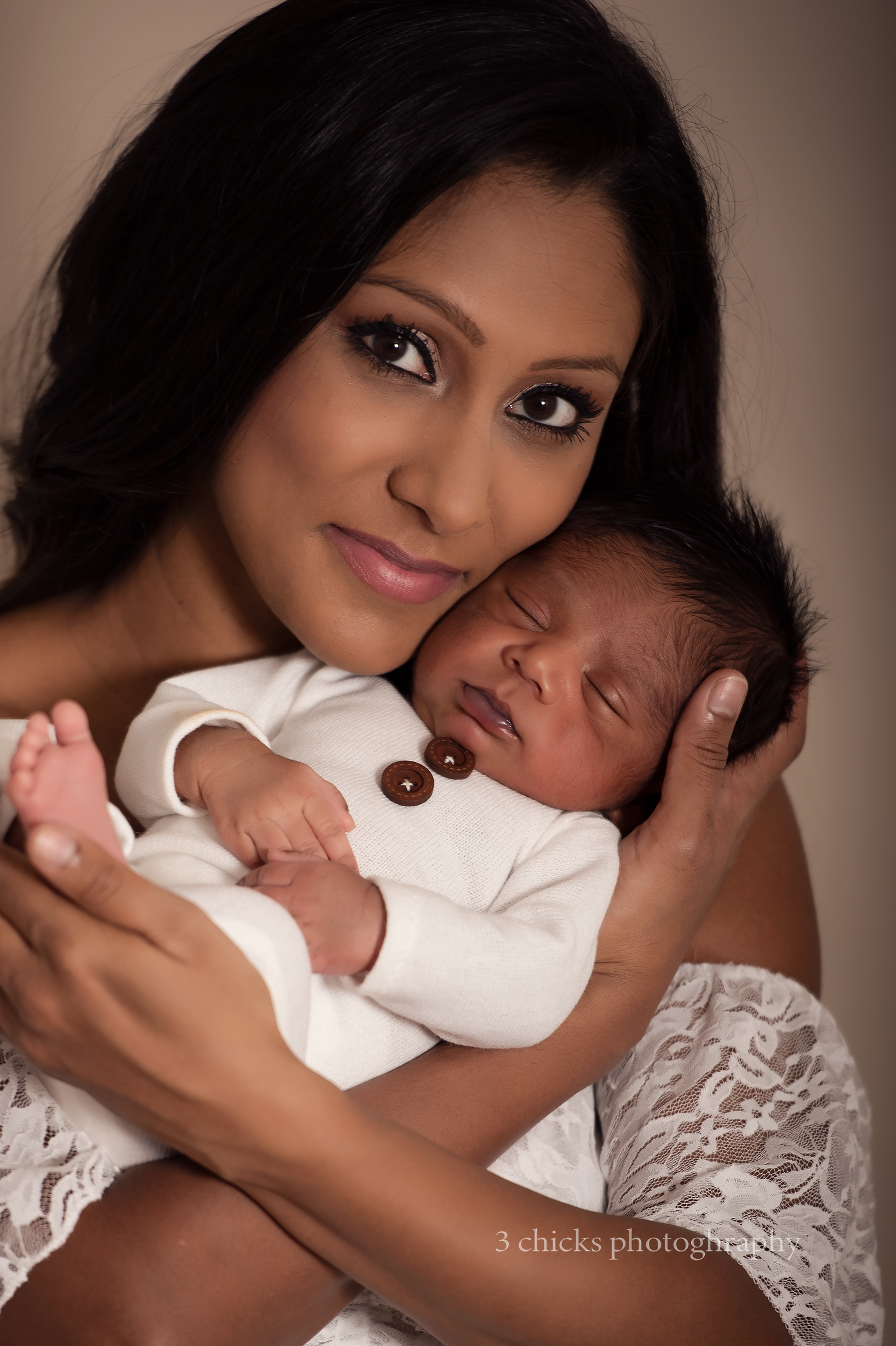 A beautiful mother tenderly holds her sleeping newborn baby dressed in white, in a professional studio portrait photo.