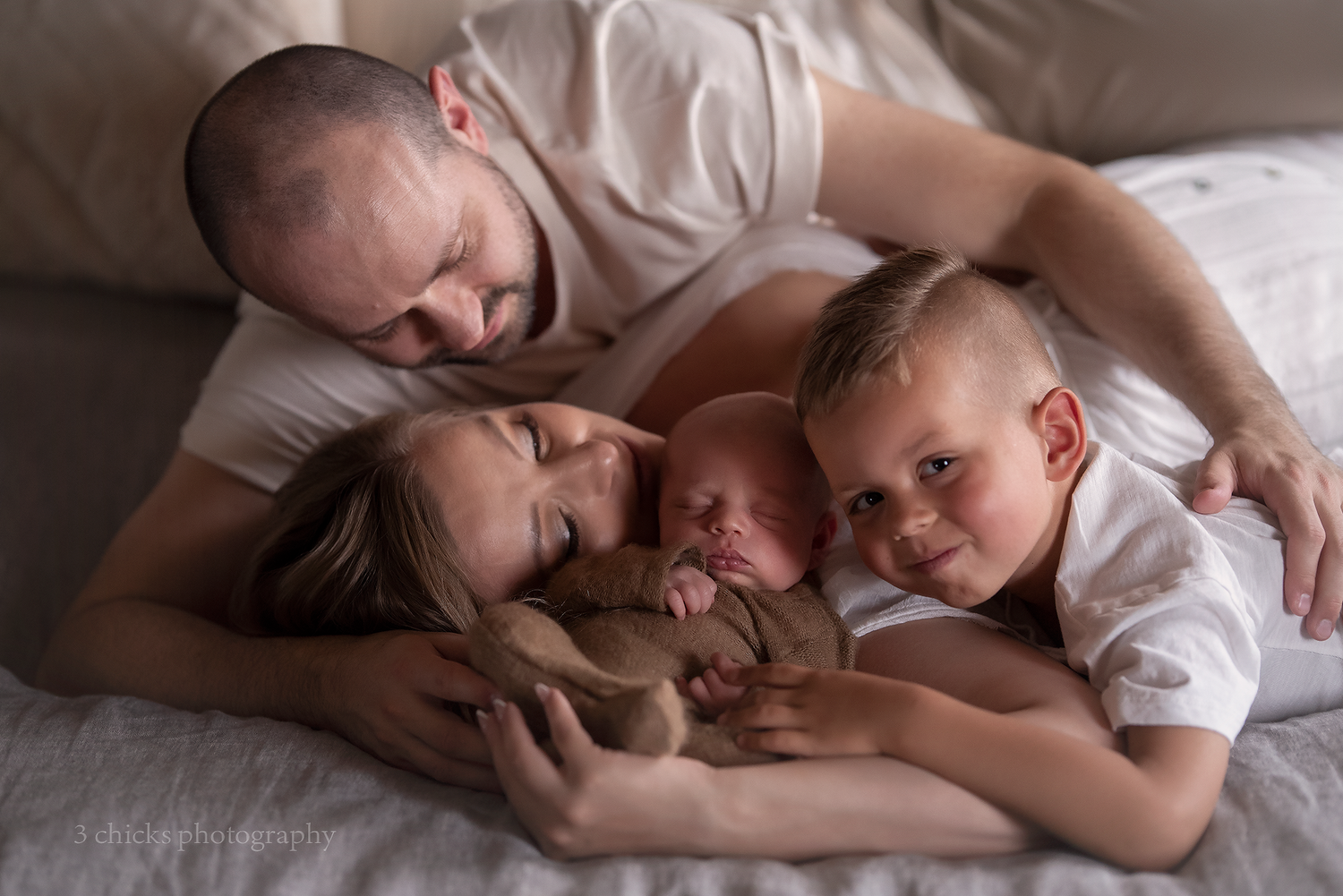 Family lying on bed together smiling with newborn baby and stuffed bear.