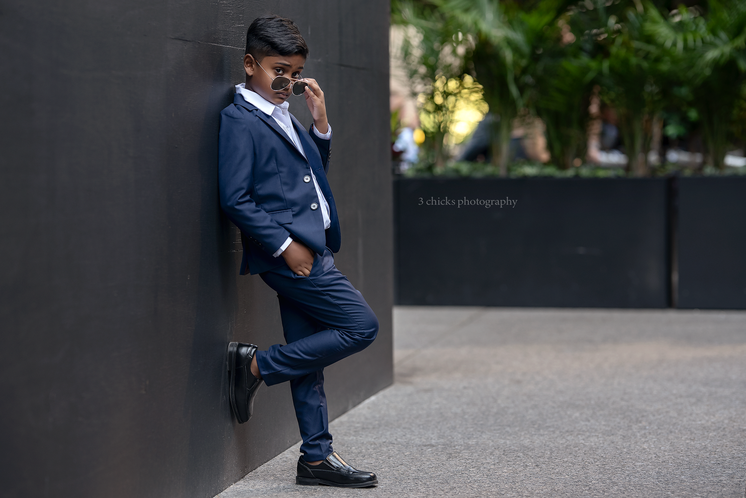 Stylish young man in navy suit leaning against dark wall while talking on phone outdoors.
