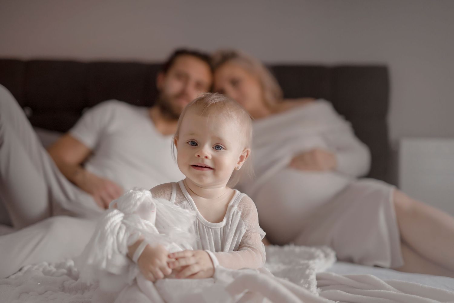 Baby in white onesie sitting on bed with parents resting in background during a cozy family photoshoot.