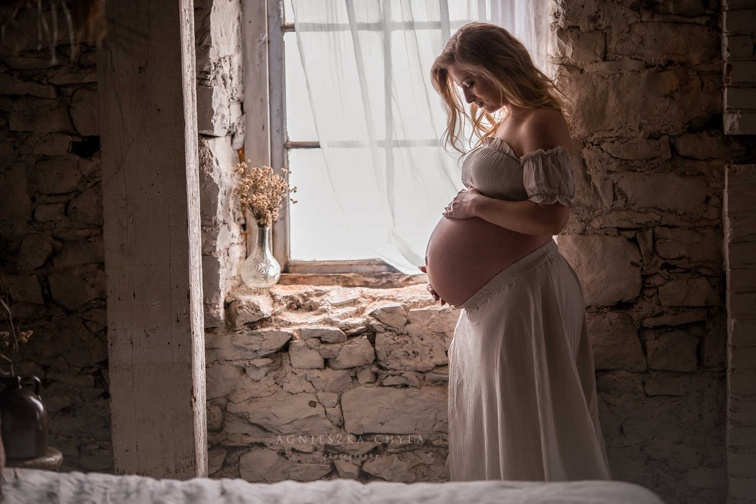 Pregnant woman in white dress cradling baby bump by rustic stone window in soft light.