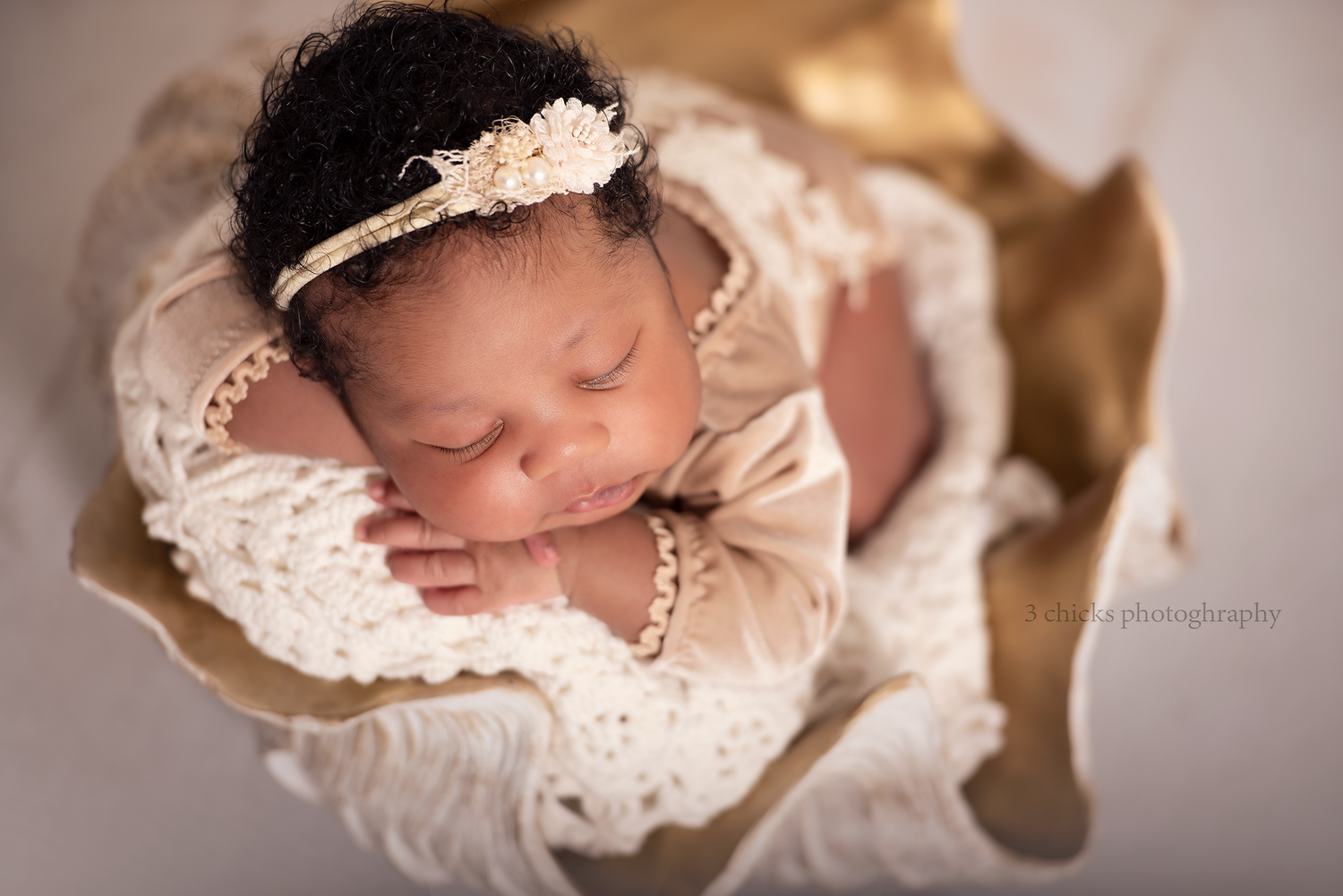 Sleeping newborn baby wrapped in ivory lace with a floral headband, nestled in a golden bowl for a professional photo.