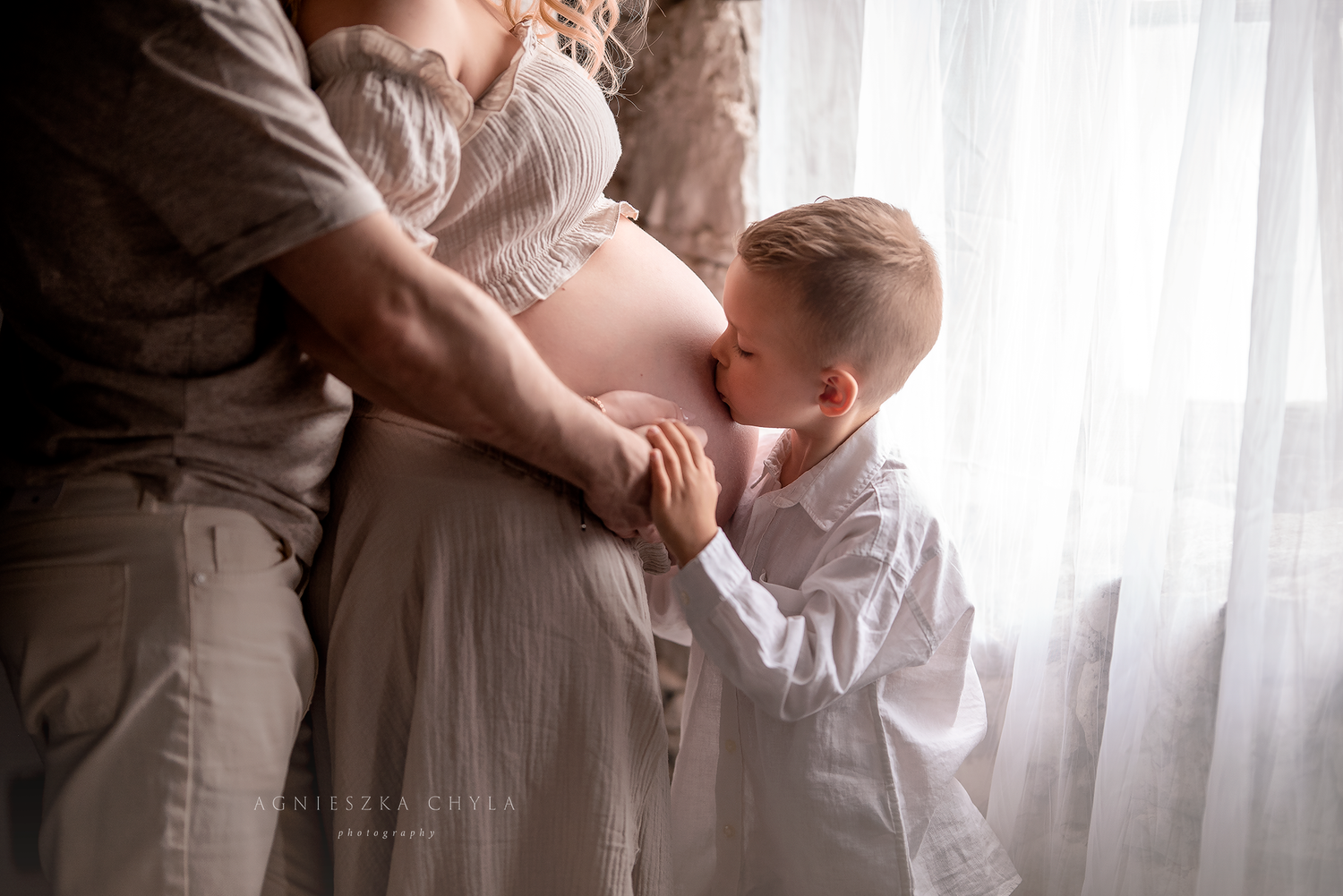 A young boy kisses his pregnant mother's belly in a soft, natural light maternity photo session.