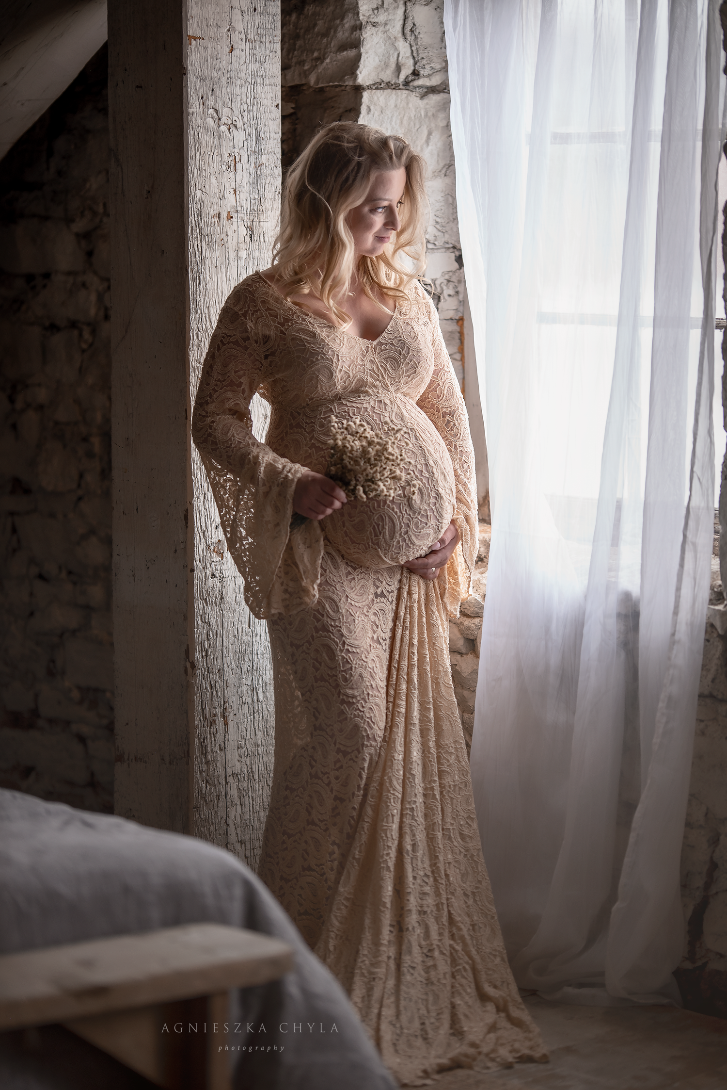 Pregnant woman in elegant beige lace gown holding baby's breath flowers, posing by a window in soft natural light.