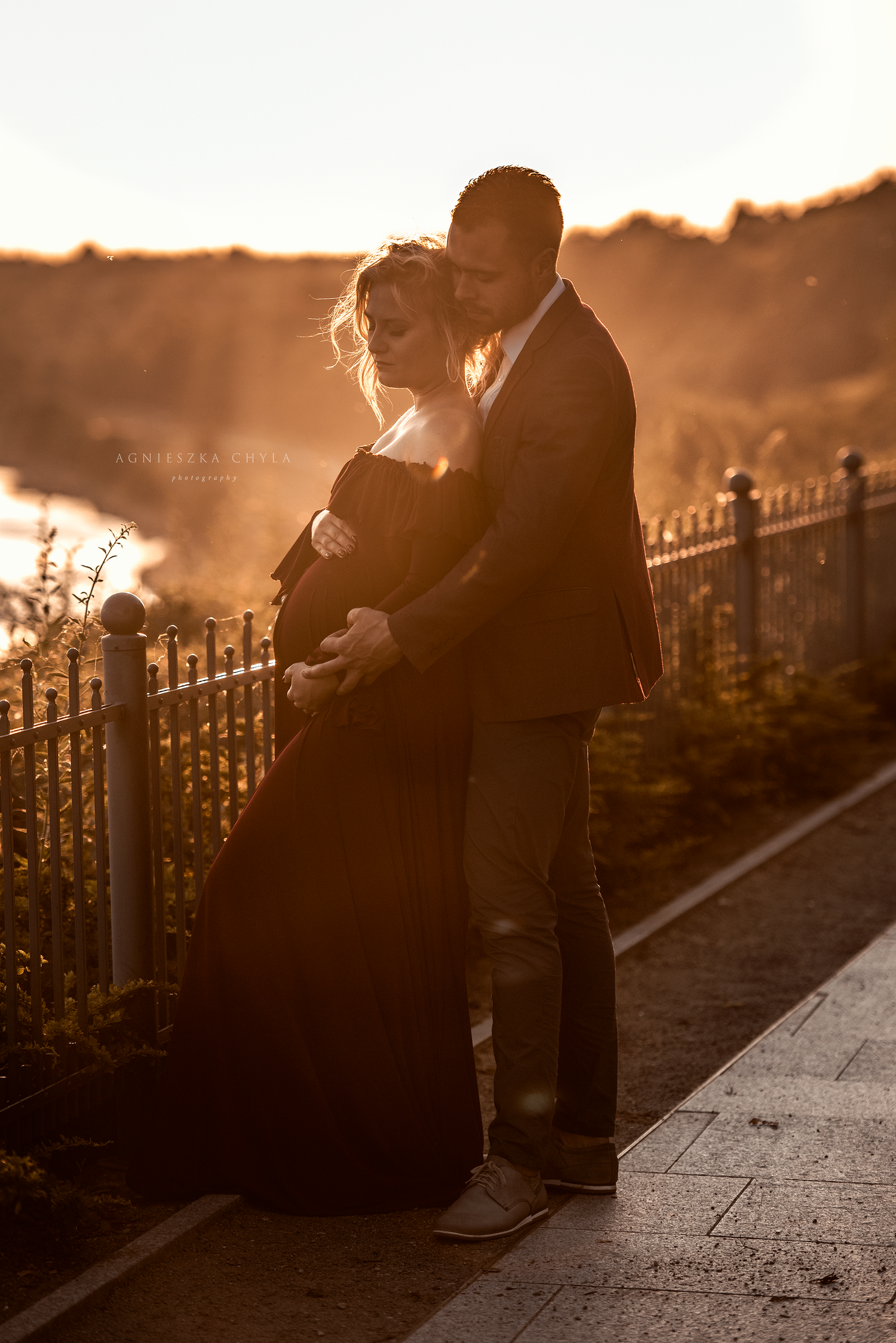 Couple embracing at golden hour sunset on a scenic walkway, woman in black maternity gown, warm backlit glow.