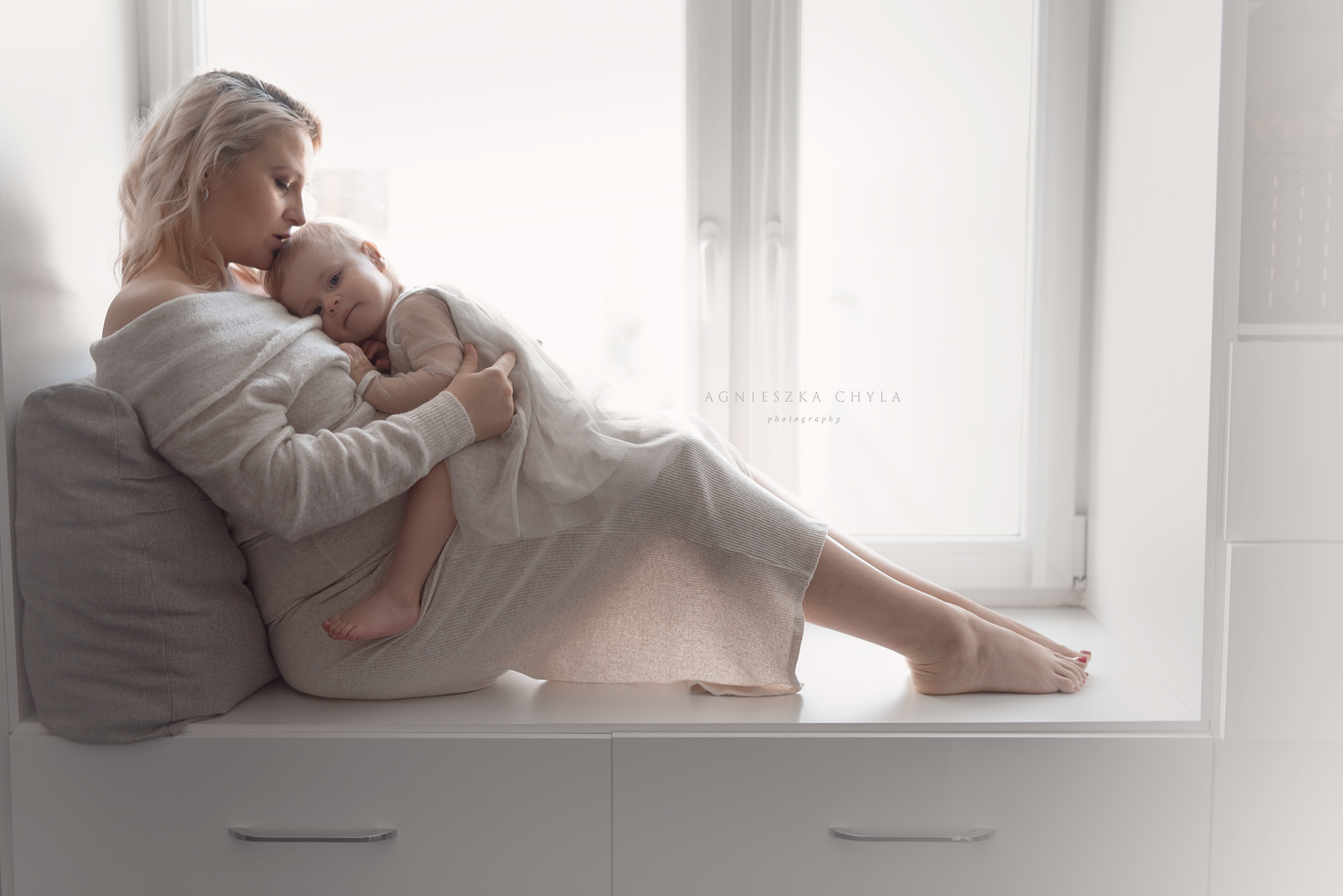 A mother in a white robe tenderly holds her sleeping baby by a bright window on a cushioned seat.