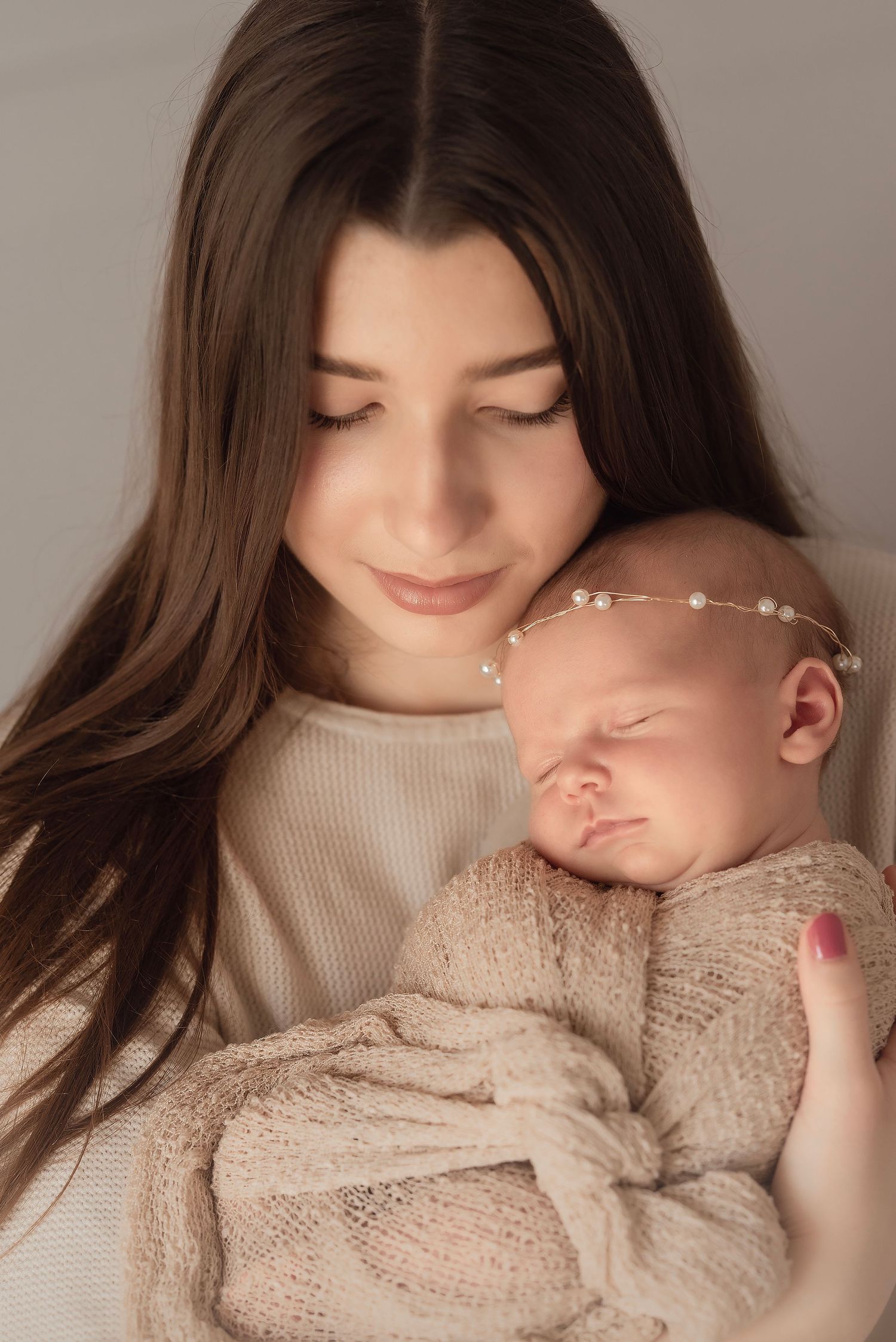 A young mother with long dark hair tenderly holds her sleeping newborn baby wrapped in a soft beige knit blanket.