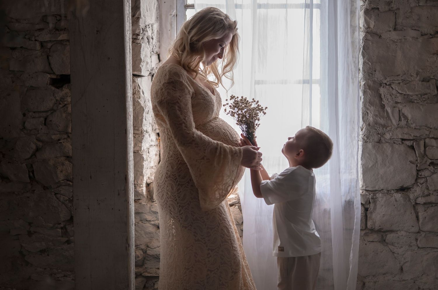 Pregnant woman in lace dress receiving flowers from toddler child by bright window indoors.