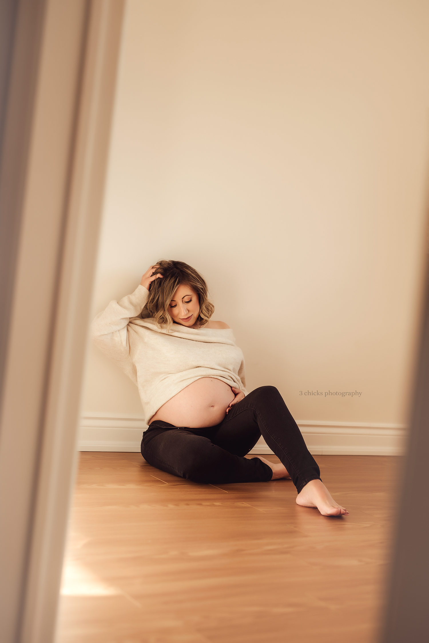 Pregnant woman sitting on hardwood floor against white wall, wearing cream sweater , cradling baby bump. Pregnancy session brampton