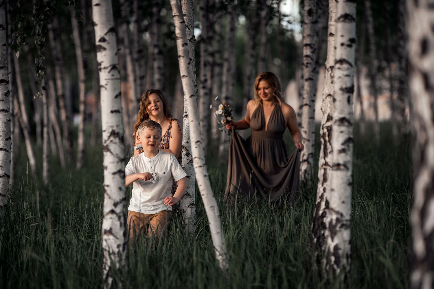 Two women and a young boy walking through birch forest holding sparklers in summer.