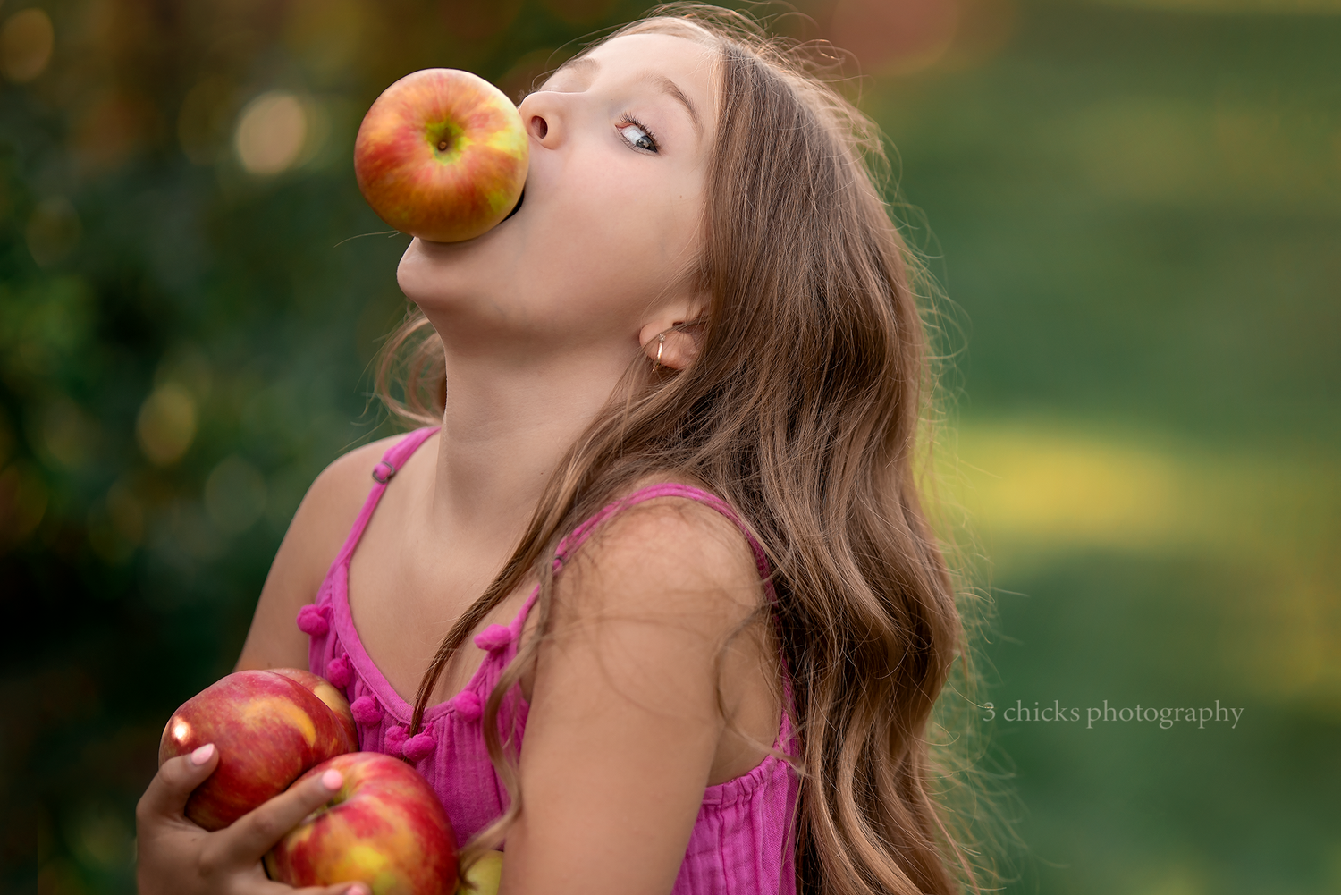 Young girl balancing a red apple on her mouth while holding more apples outdoors.