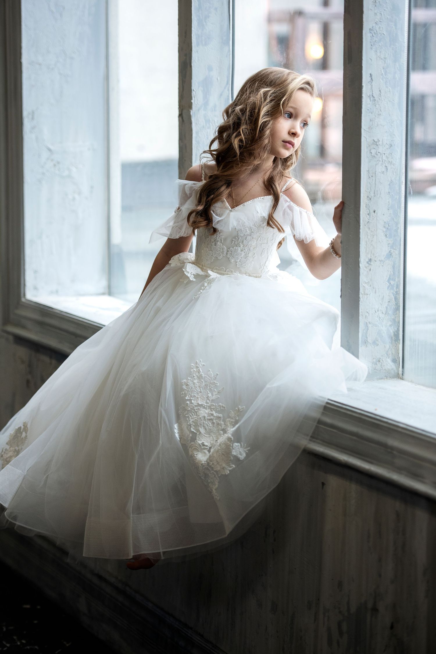A young girl in a white off-shoulder ball gown sits gracefully on a window ledge.