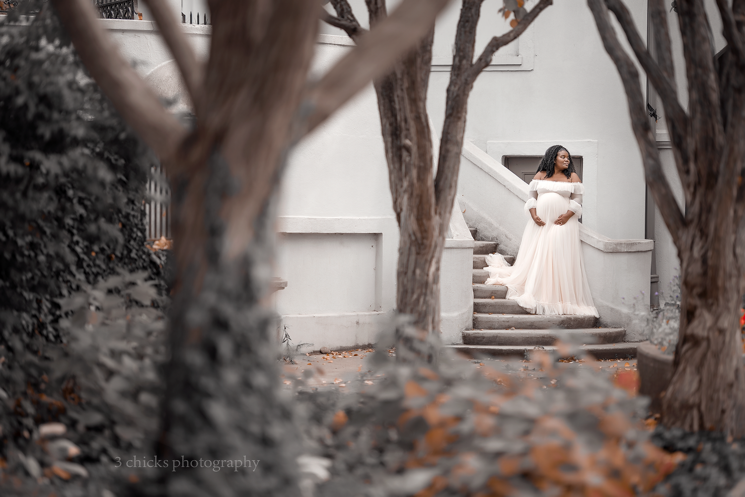 Pregnant woman in white off-shoulder gown poses gracefully on outdoor stone steps surrounded by bare trees.
