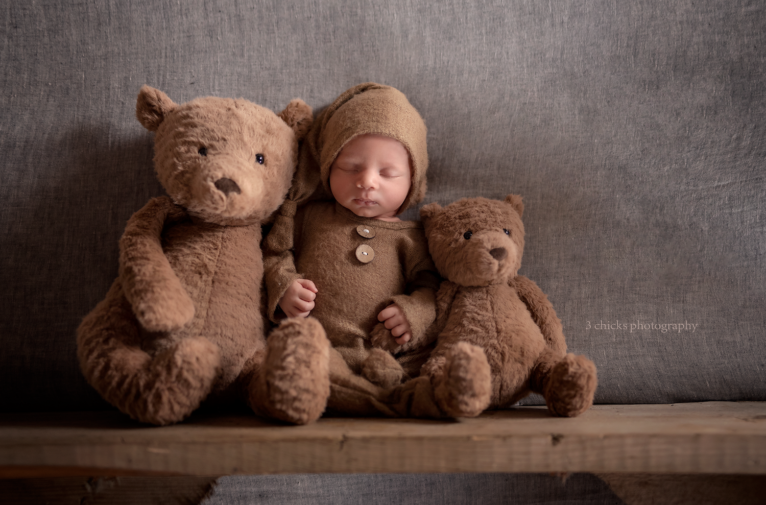 Sleeping newborn baby in brown bear outfit posed between two teddy bears on wooden bench. Baby photography Burlington