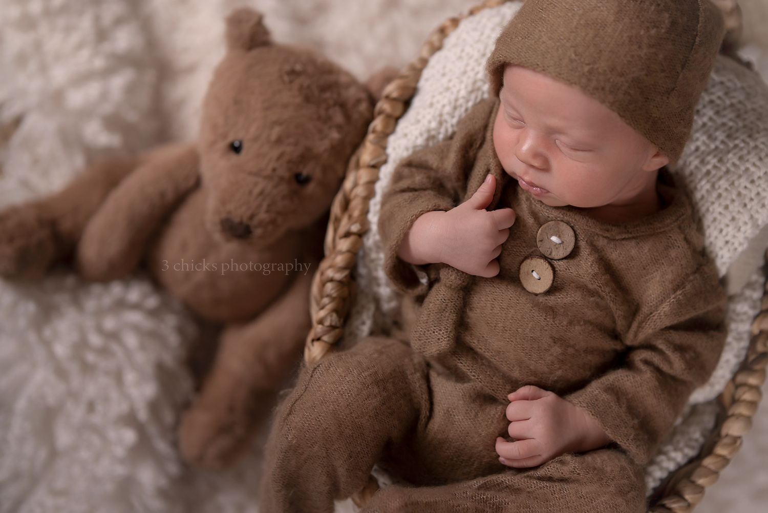 Peaceful newborn baby in brown knit outfit with buttons resting in a basket beside a teddy bear.