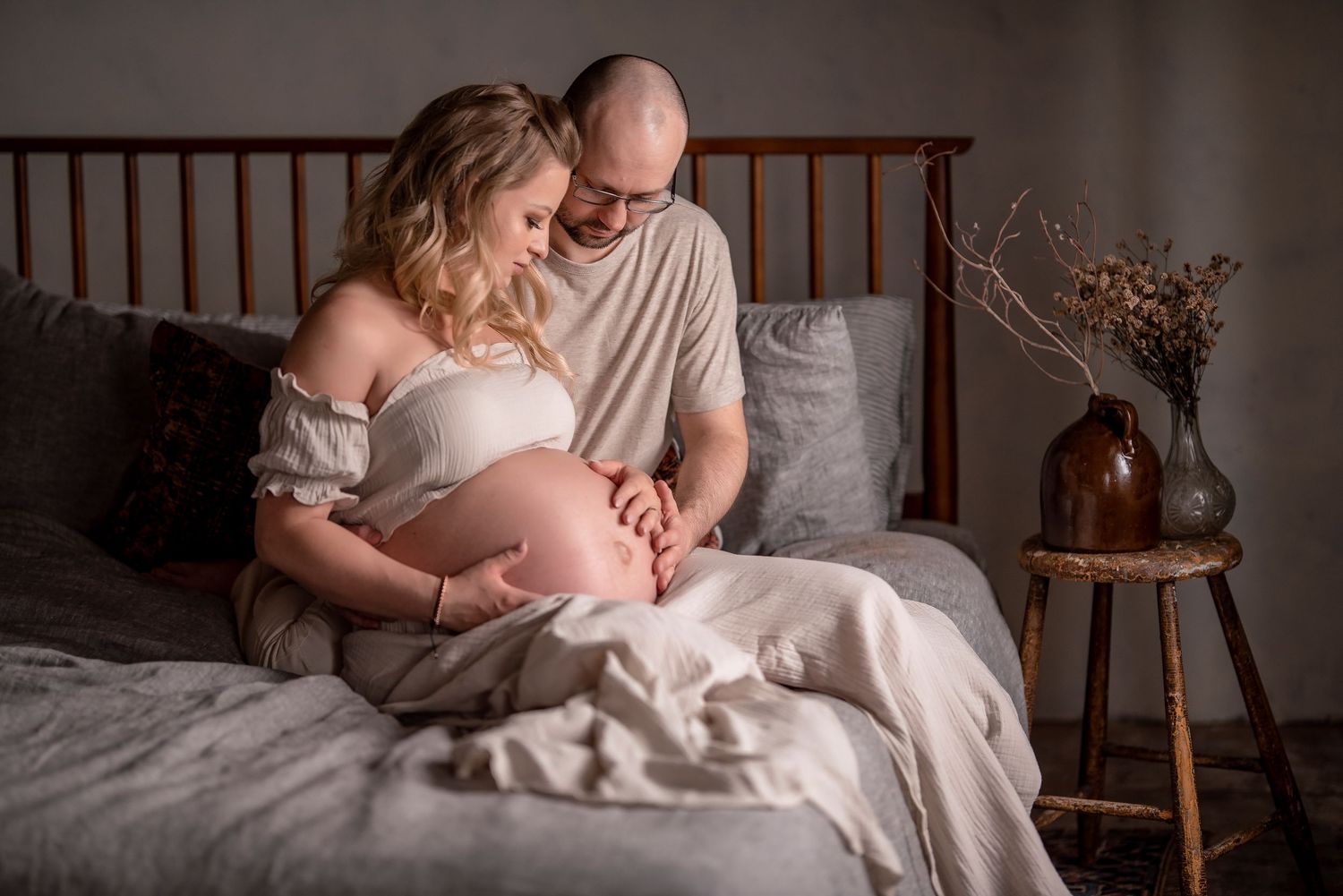 Loving couple sits on bed gazing at pregnant belly during intimate maternity photo session in cozy bedroom.